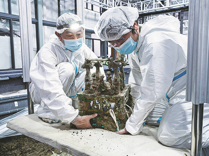 Dos arqueólogos extraen una estatua de bronce de un pozo de sacrificio, 12 de junio del 2022. [Foto: Yu Jia/China Daily]