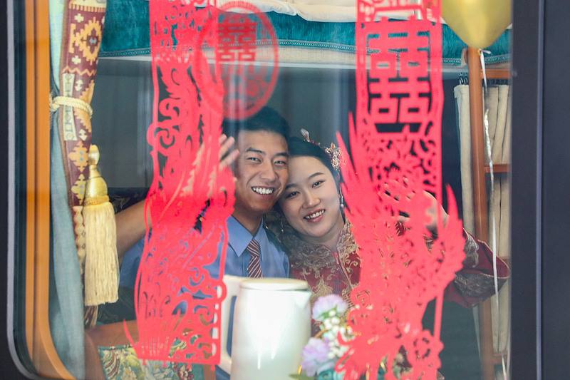Una pareja de recién casados saluda desde la ventana mientras viaja en tren hacia el lago Salado de Chaka, el 19 de agosto de 2026. Con motivo del Festival Qixi de China, 26 parejas —todas empleadas ferroviarias— celebraron una boda colectiva en la plaza frente a la estación de tren de Xining, en la provincia de Qinghai, en el noroeste del país. Foto: VCG