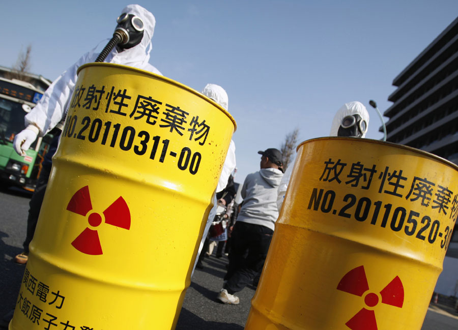 Los manifestantes antinucleares con trajes protectores de radiación empuje barriles con etiquetas de residuos radiactivos procedentes de las plantas de energía nuclear Kansai Electric Power Co. (izquierda) de Ohi y Tokio Electric Power Co. (TEPCO) de Fukushima, durante la marcha en Tokio, el 9 de marzo de 2014. [Foto/agencias]